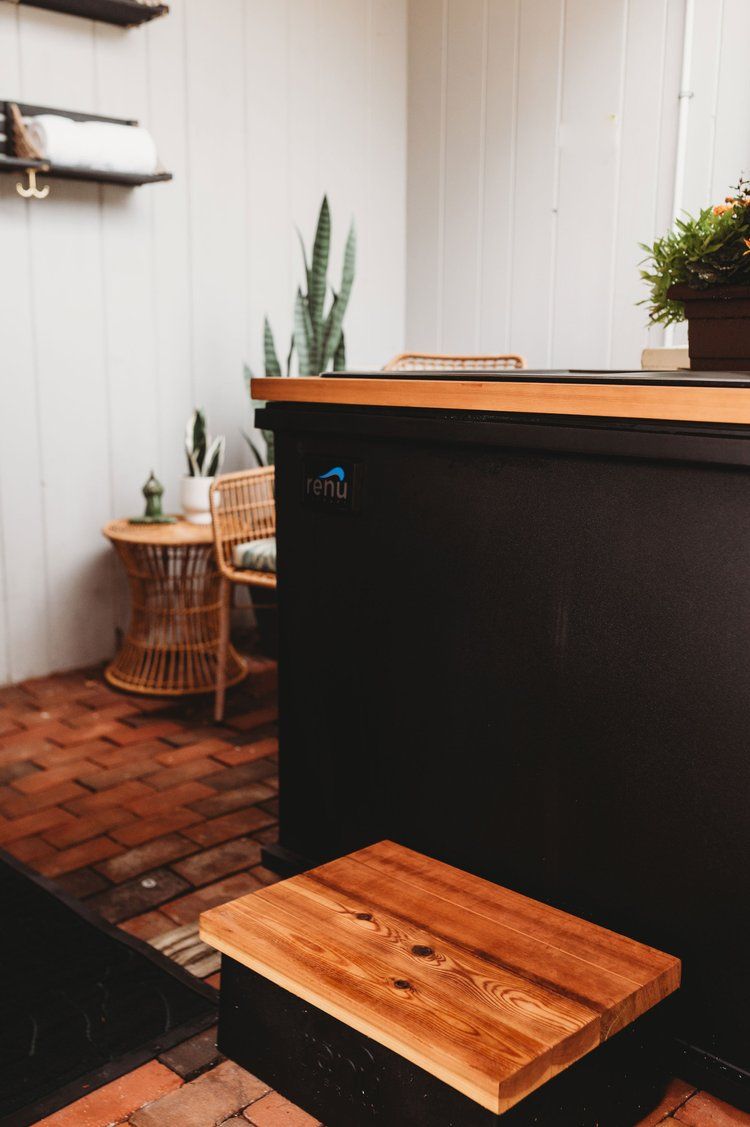 Black hot tub with wooden step in a brick-floored outdoor space, plants, and wicker furniture.