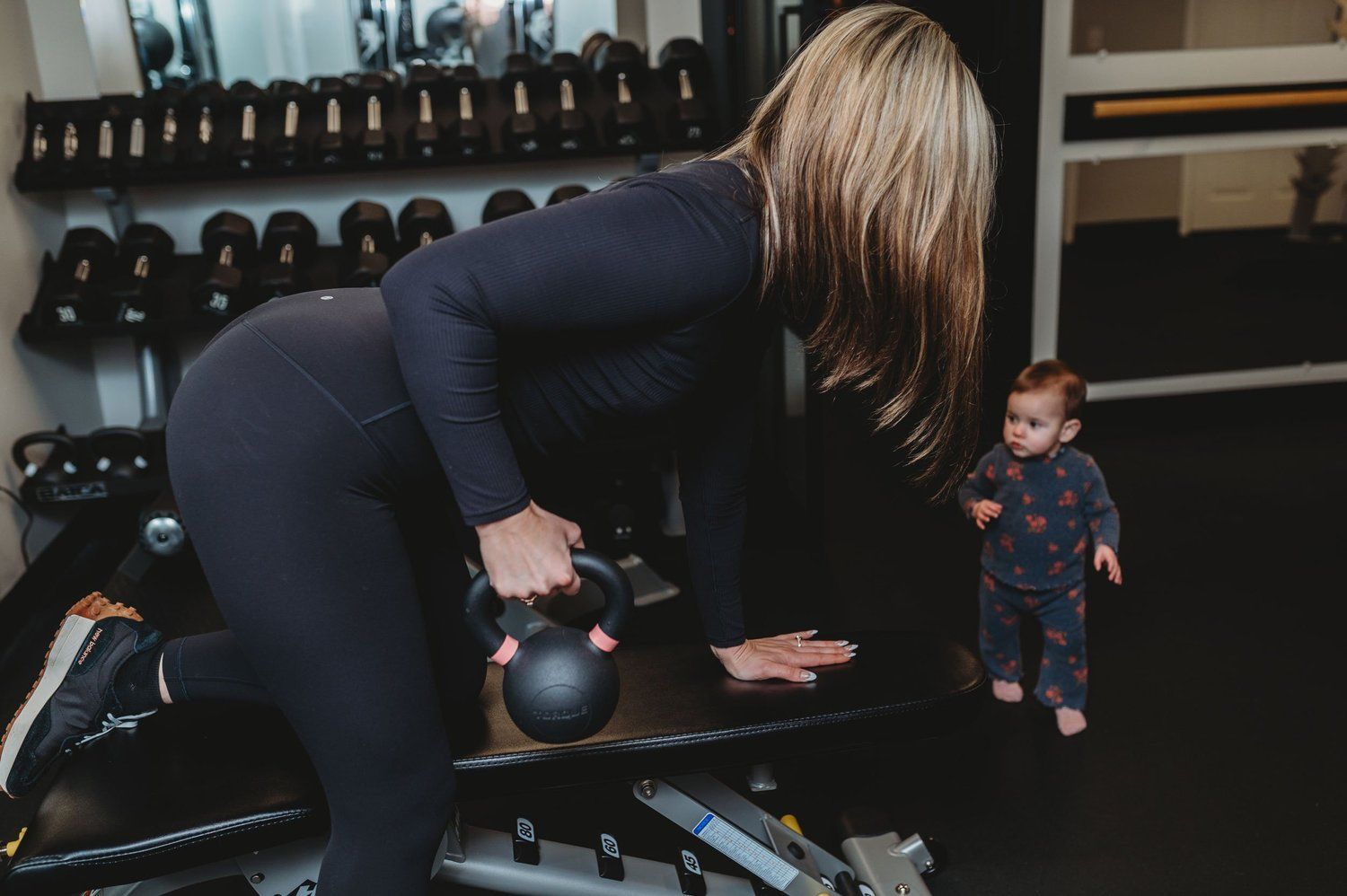 Woman in gym lifting kettlebell; child watches nearby.
