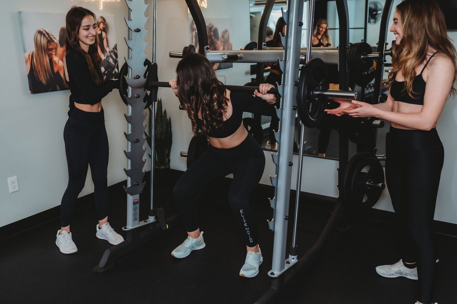 Three people in a gym; one doing a squat with assistance. They wear athletic clothes.