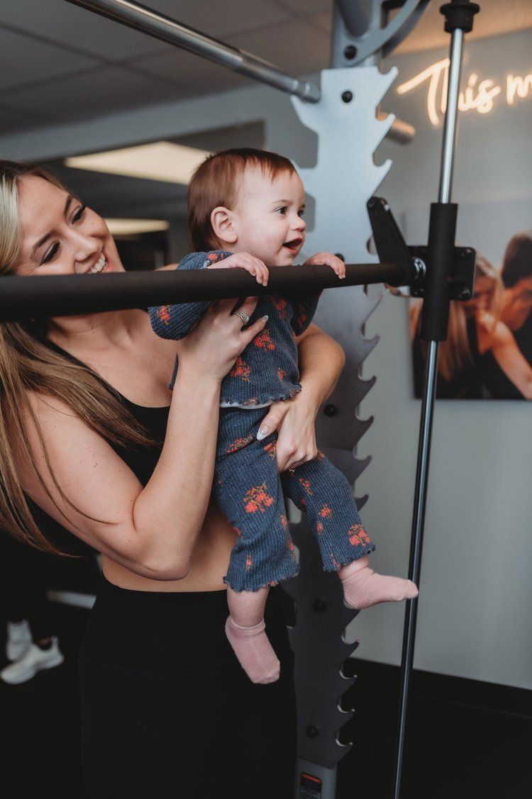 Woman holding baby up to a gym machine bar; baby smiles and looks to the side.