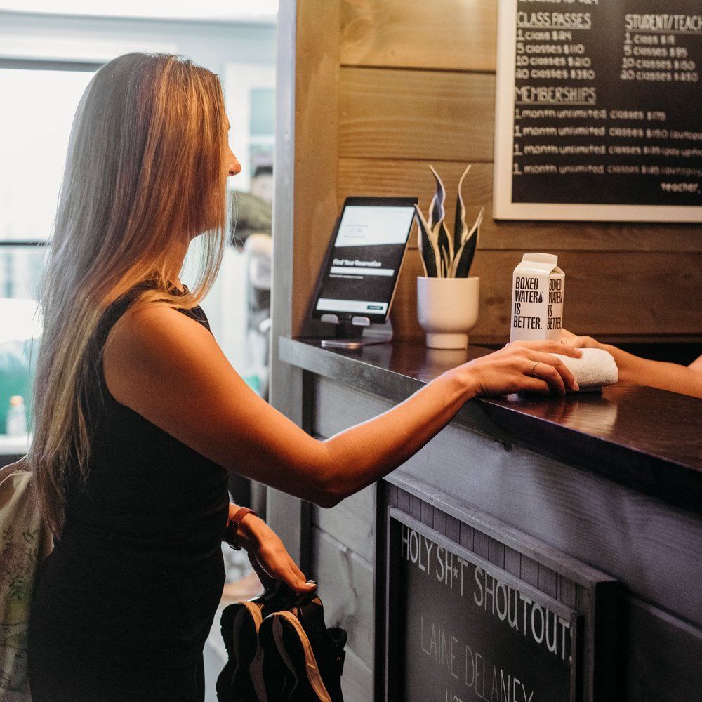 Woman at a counter with a tablet and plant, handing something to another person.