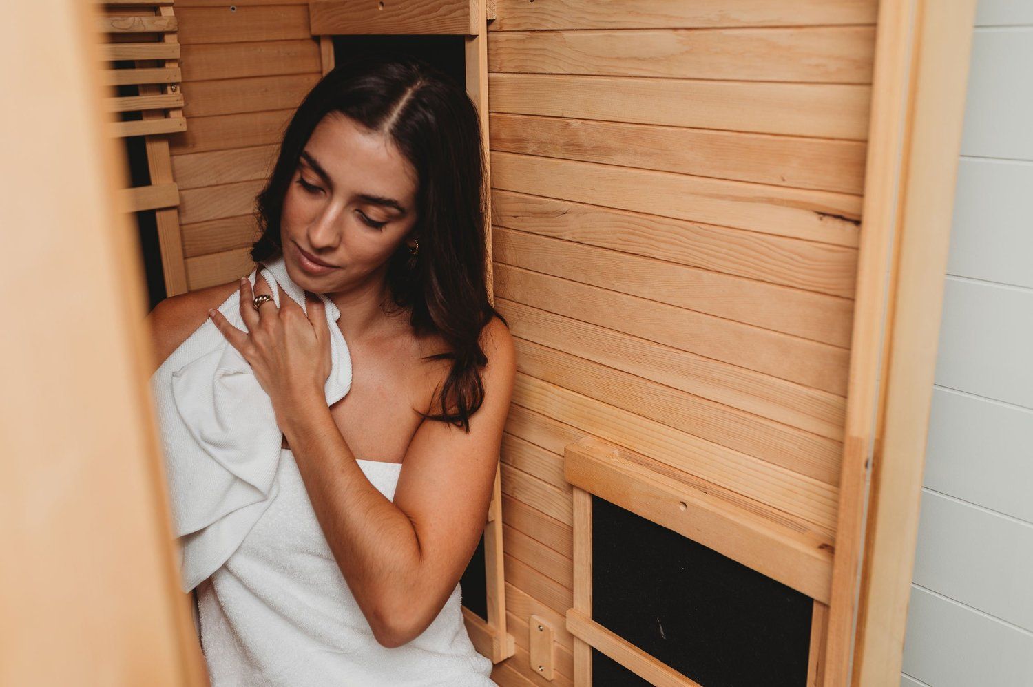 Woman in a wooden sauna, wrapped in a white towel, wipes her shoulder.