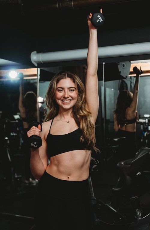 Woman in gym holding kettlebell overhead and dumbbell.