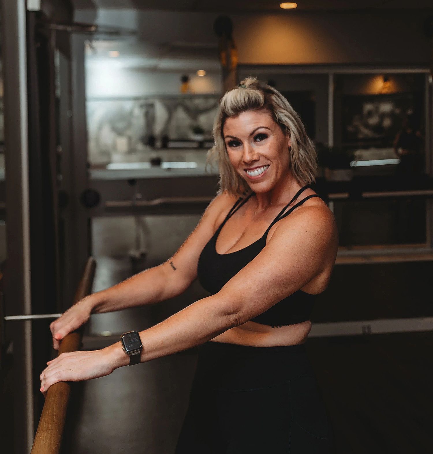 Woman in black workout clothes smiling, leaning on a barre in a studio.