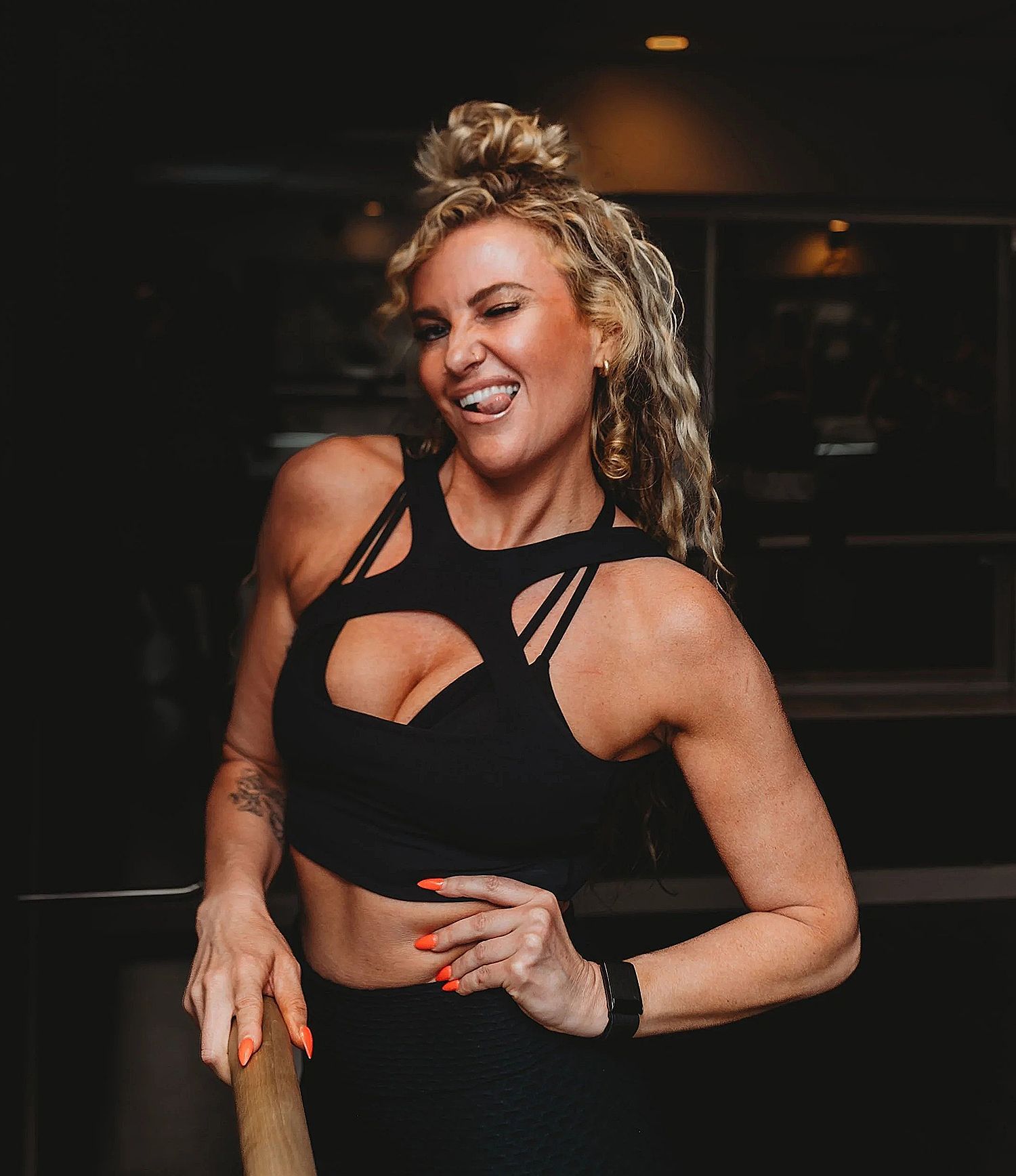Woman in a black sports bra and leggings smiles, holding a pole in a gym.
