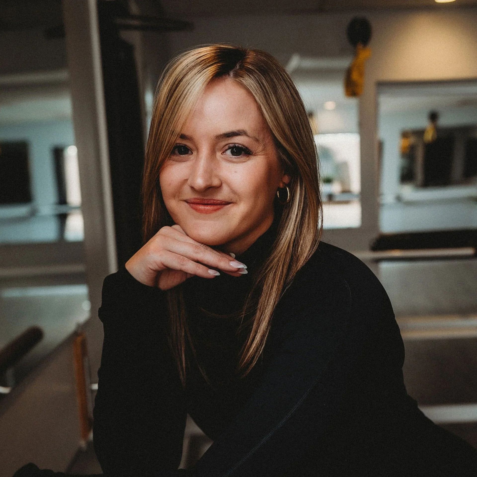 Woman with blonde hair, resting chin on hand, smiling. Wearing a black turtleneck, indoors.