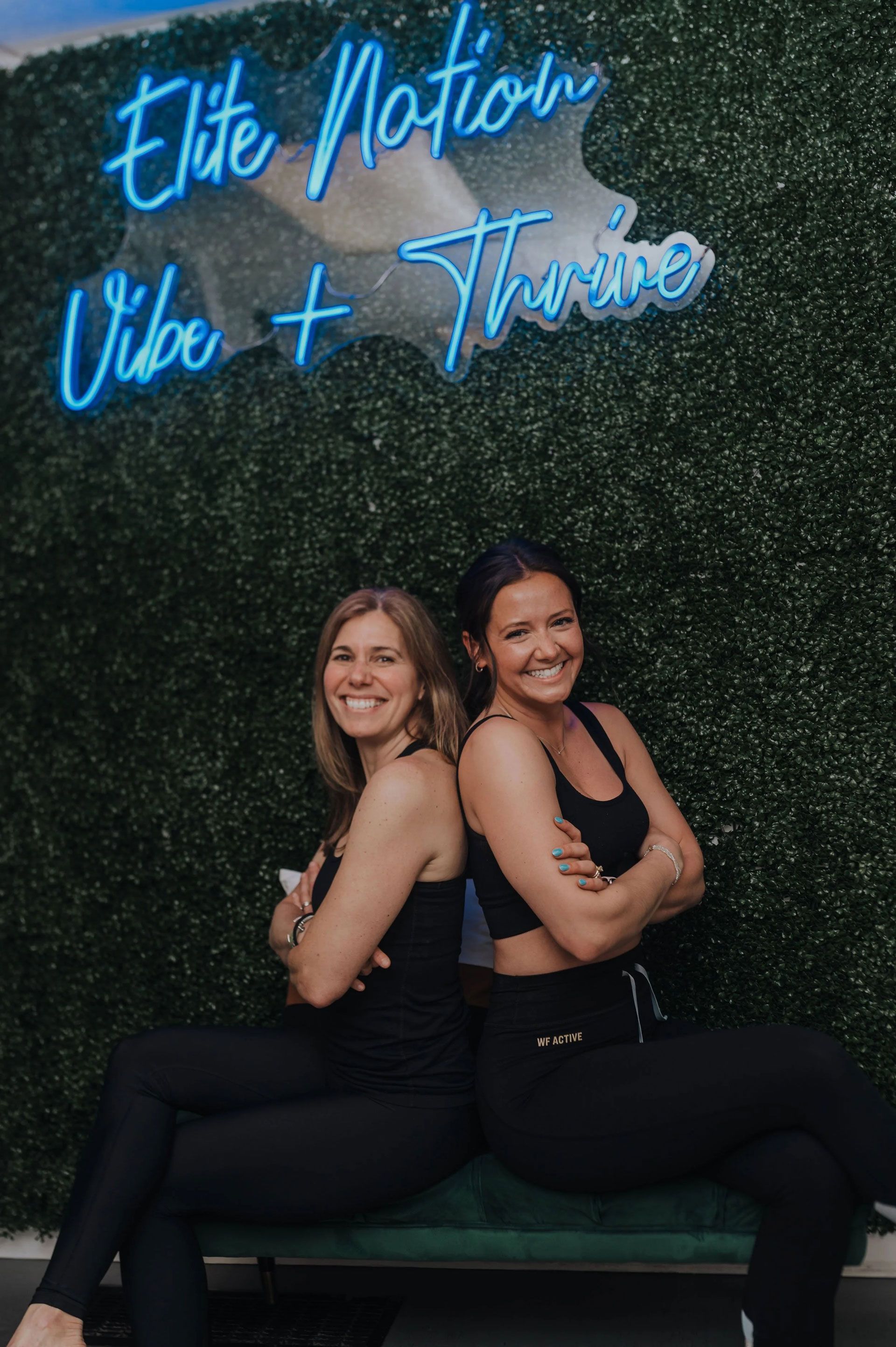 Two women in activewear smile, back-to-back, in front of a neon sign that reads "Elite Nation Vibe + Thrive."