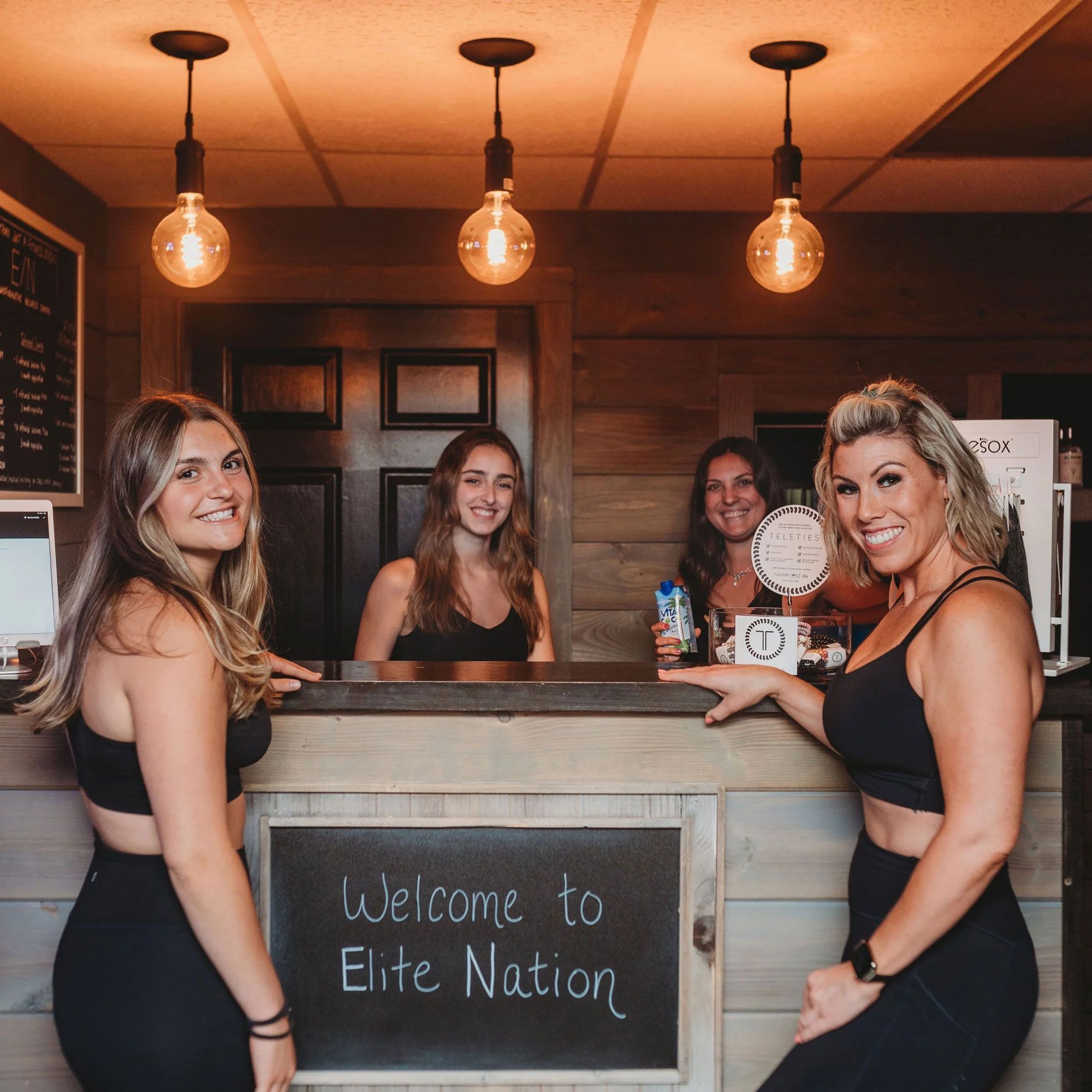 Four women at a reception desk with "Welcome to Elite Nation" on it. Rustic interior, Edison bulb lights.