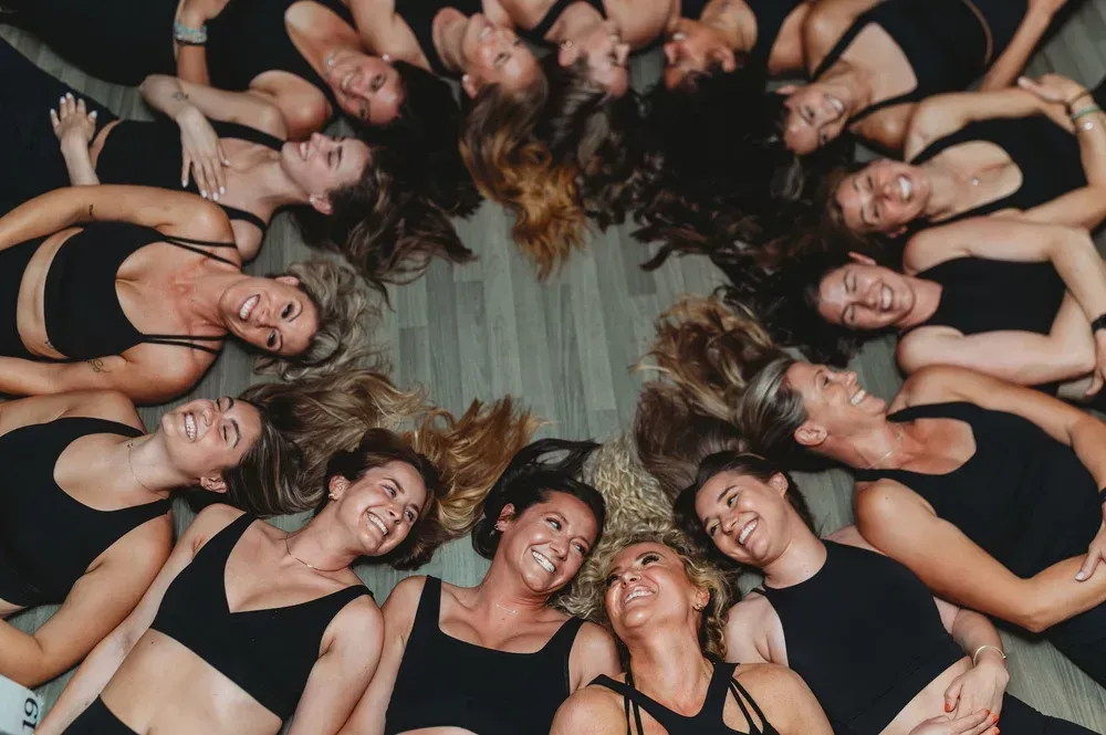 A circle of smiling people in black activewear lying on a light-colored floor, heads together.