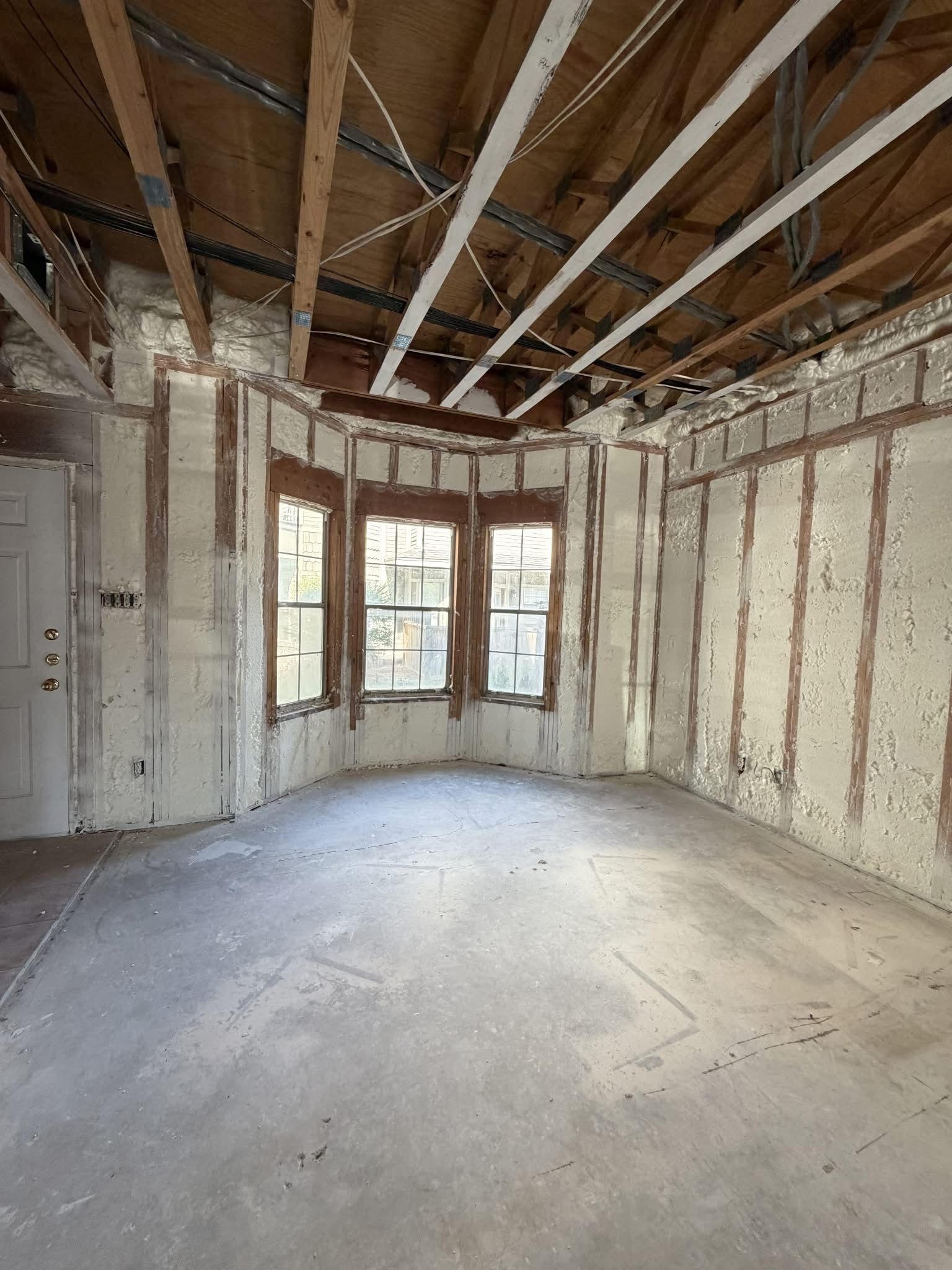 Unfinished room with exposed ceiling beams, bare walls, and three front windows with daylight.
