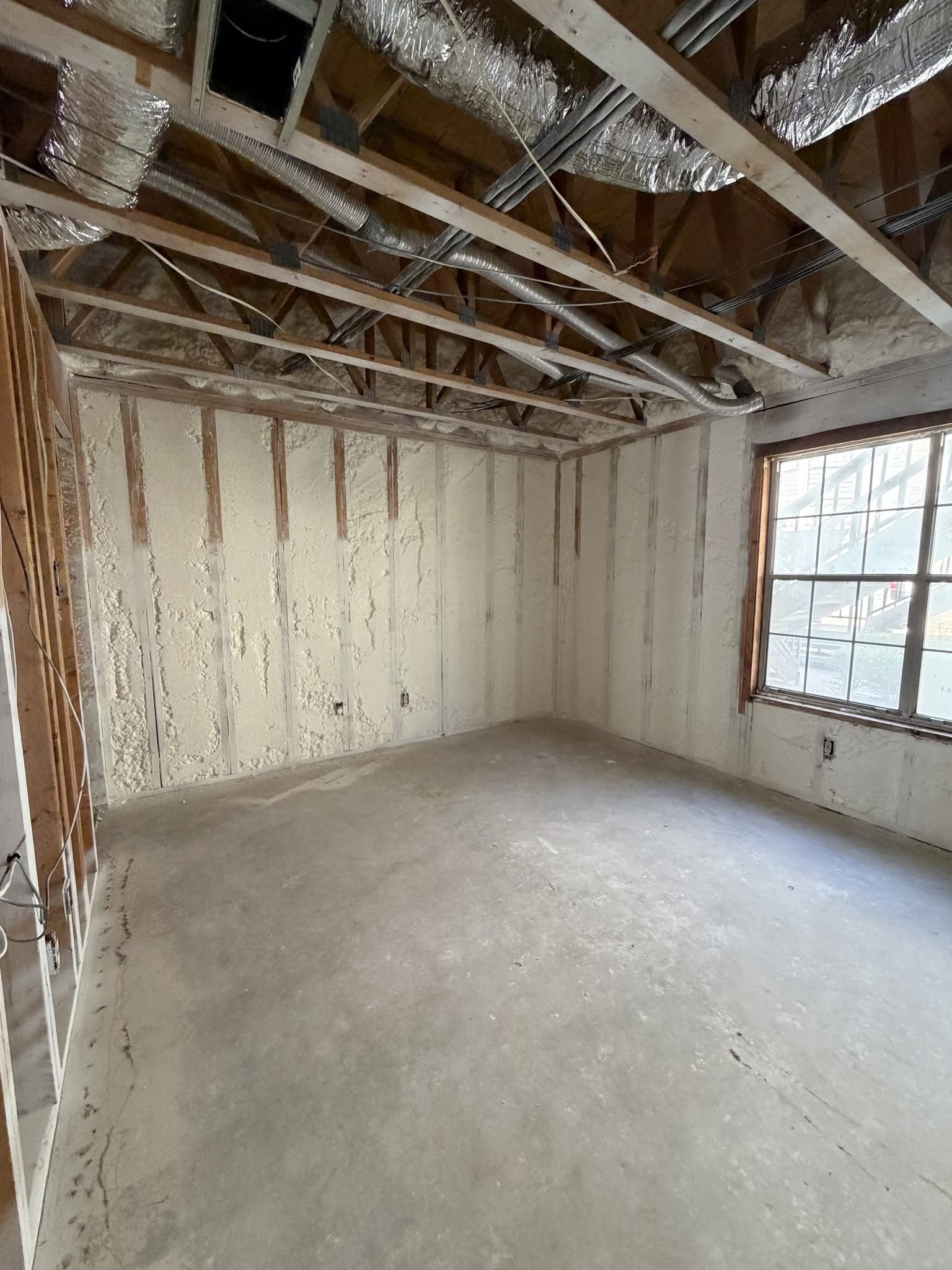Bare unfinished room with exposed ceiling beams, insulation, and a window on the right.