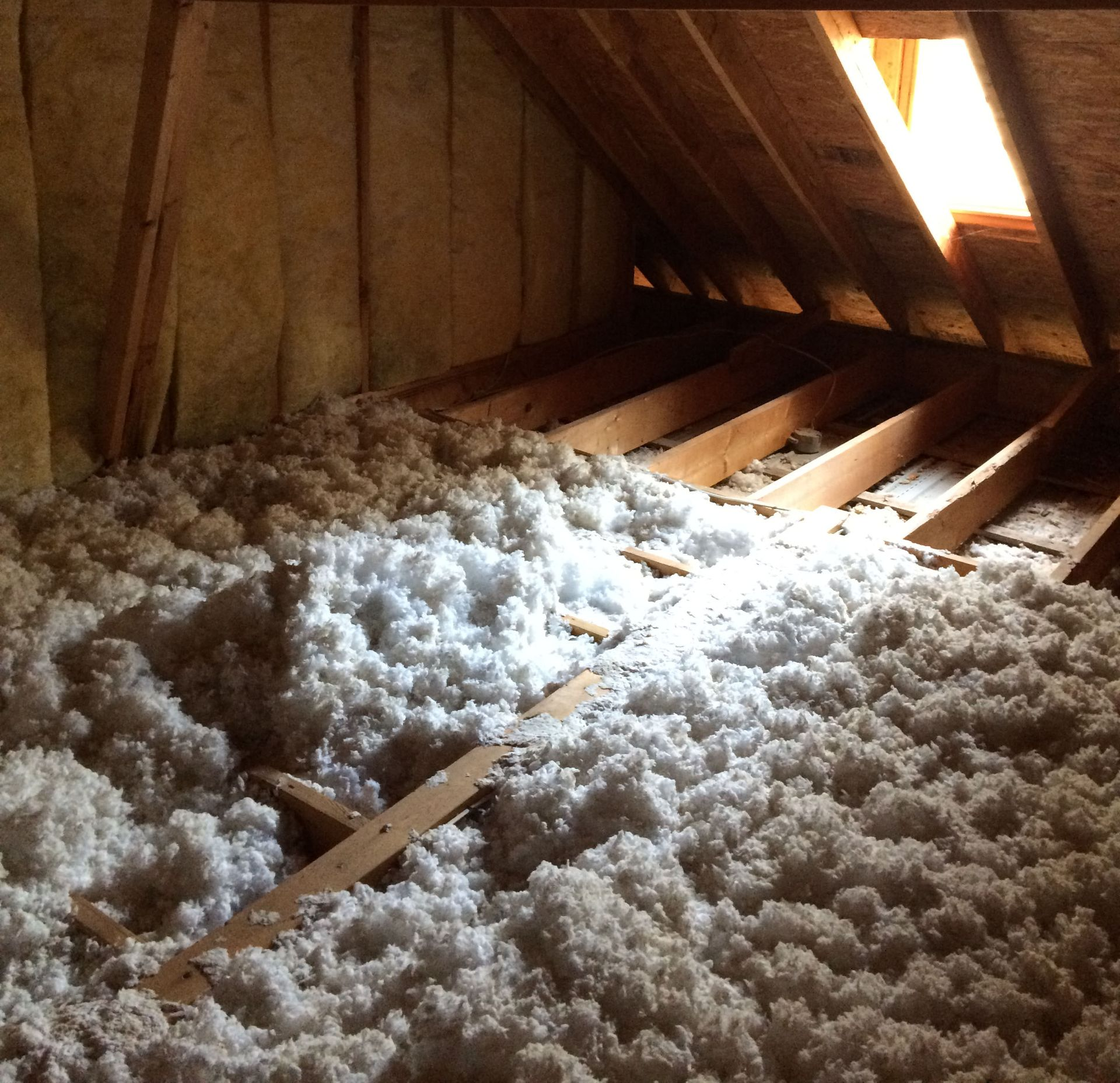 Attic with thick white insulation between wooden rafters, lit by a small window.