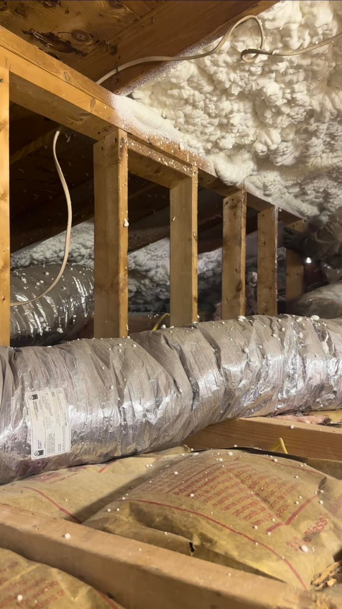 Attic view with wooden framing, spray foam insulation, and HVAC ductwork wrapped in foil.