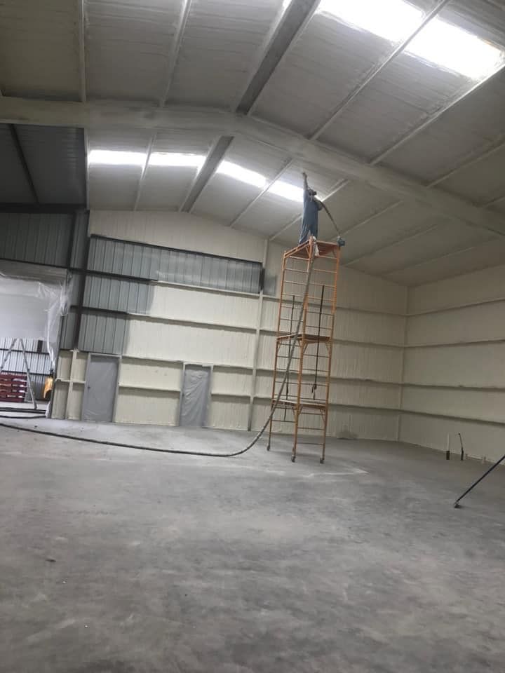 Man spraying insulation on a warehouse ceiling from a tall ladder. Walls are partially insulated.