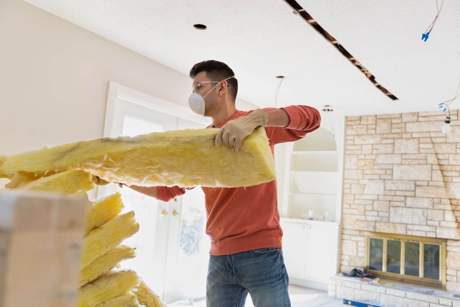Man installing insulation in a room with fireplace, wearing a mask and gloves.