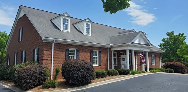 A one-story red brick building with a gabled roof, dormer windows, and a pillared porch entry, surrounded by landscaping.