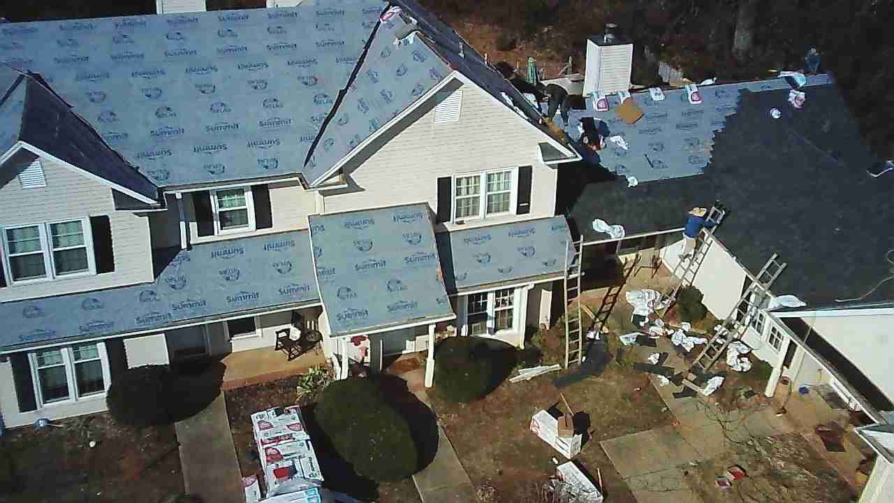 Aerial view of a two-story house under construction, featuring half-shingled gray roofing and exposed roofing underlayment.