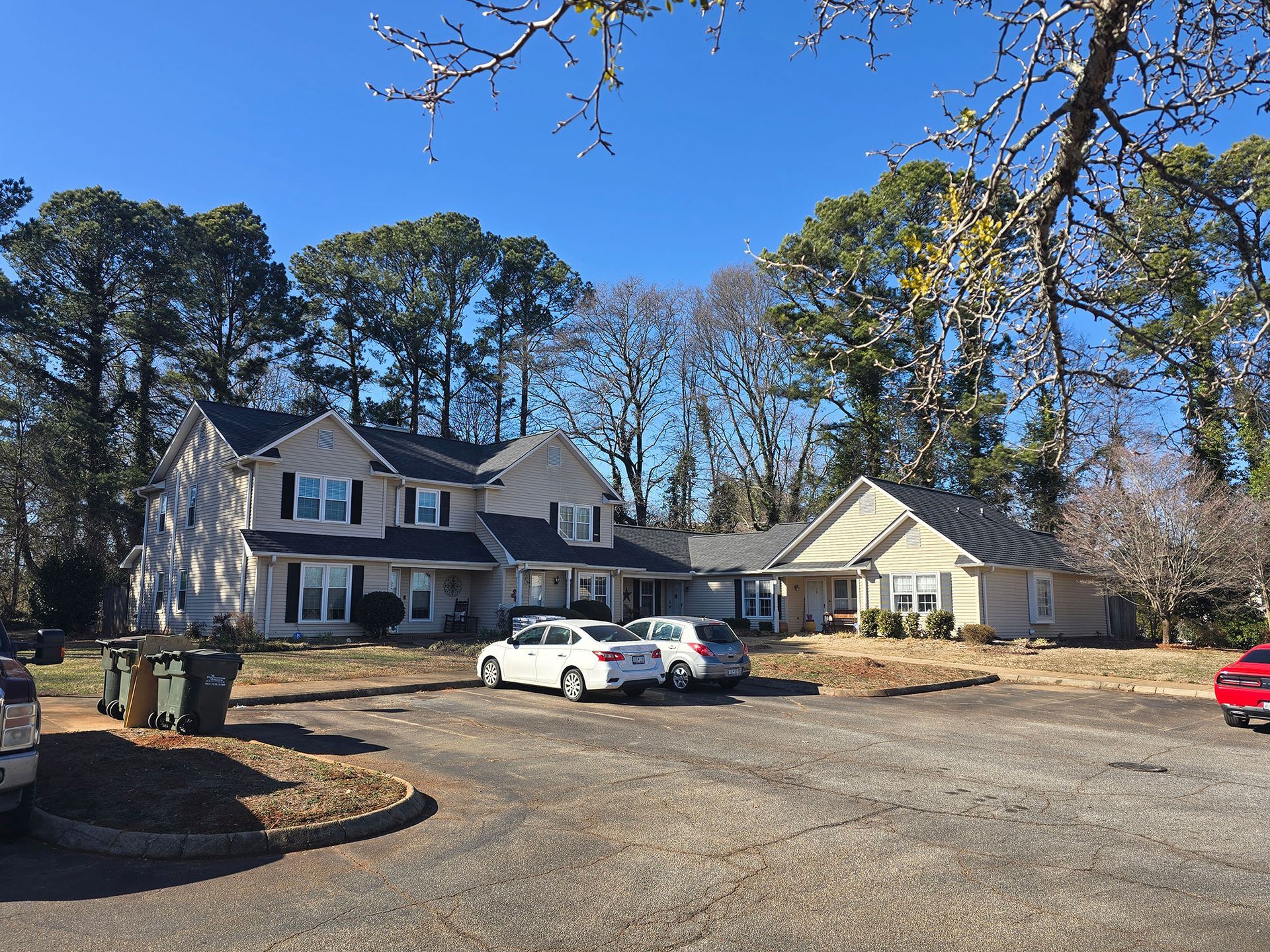 A two-story beige house and adjacent one-story structure set behind a gravel parking lot under a clear blue sky.