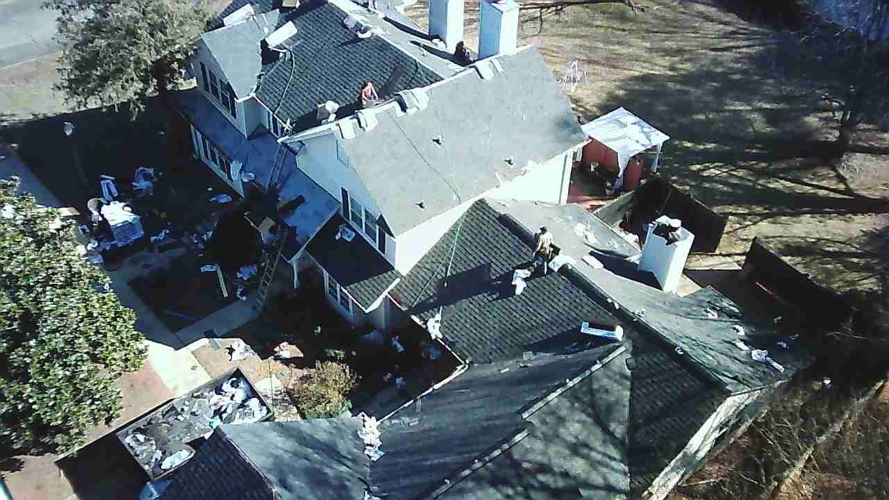 Aerial view of a residential house roof with some areas under repair or construction, surrounded by a yard and trees.