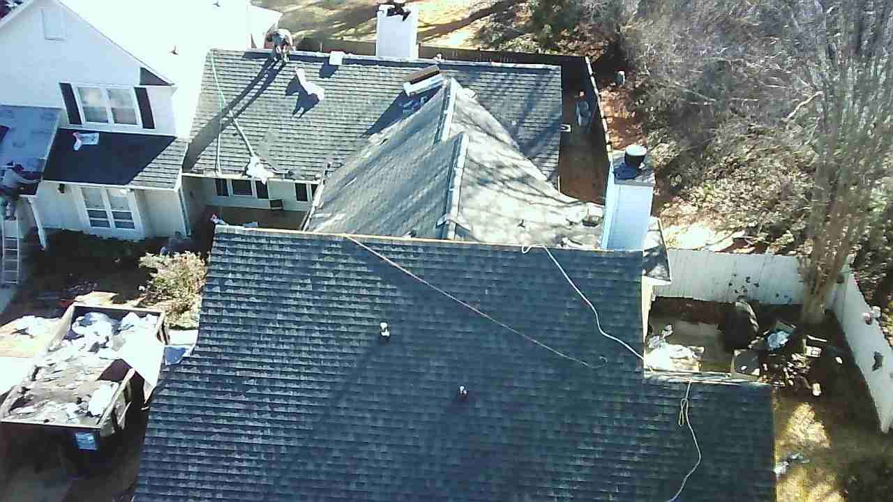An aerial view of a house undergoing a roof replacement with shingle bundles and equipment visible on and around the roof.