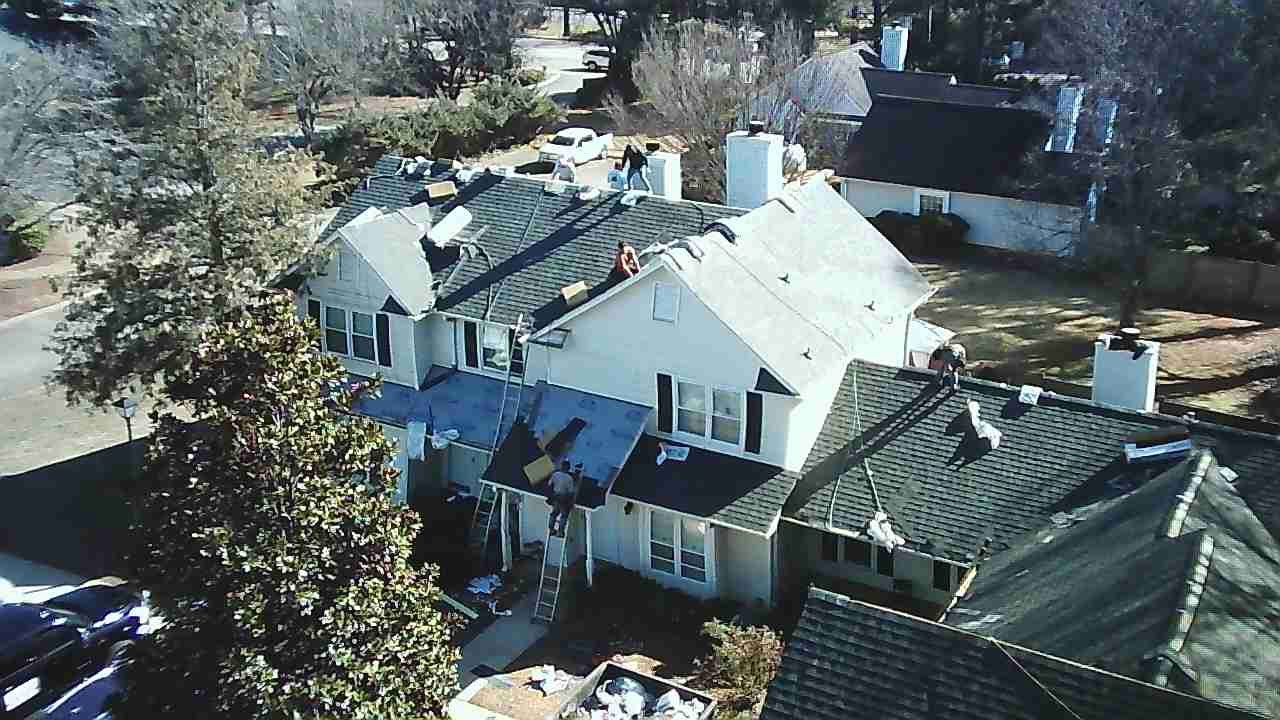 An aerial view of a two-story residential house undergoing roof repairs, with patches of bare roof deck and scattered tools.