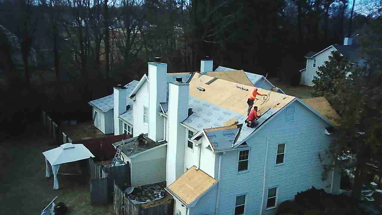 Two workers in bright orange safety gear install new roof panels on the gabled roof of a two-story residential house.