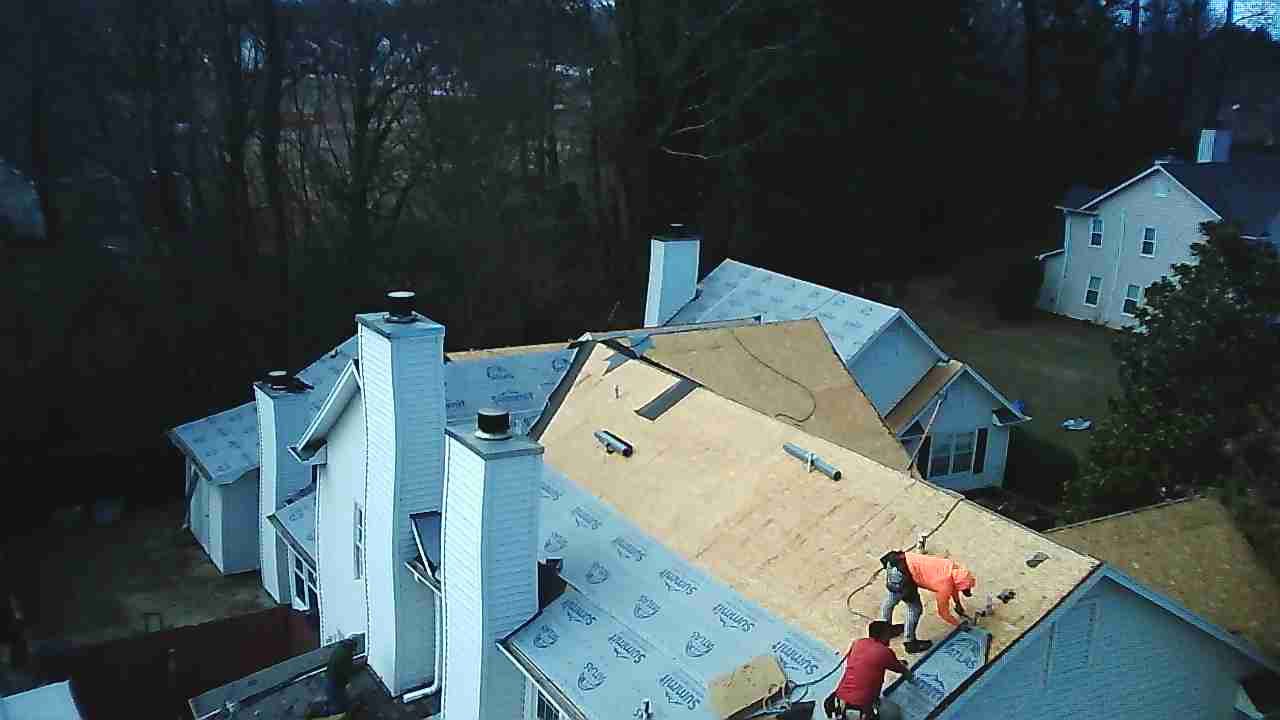 Two people work on repairing a residential roof, installing underlayment and plywood decking on a sunny day.