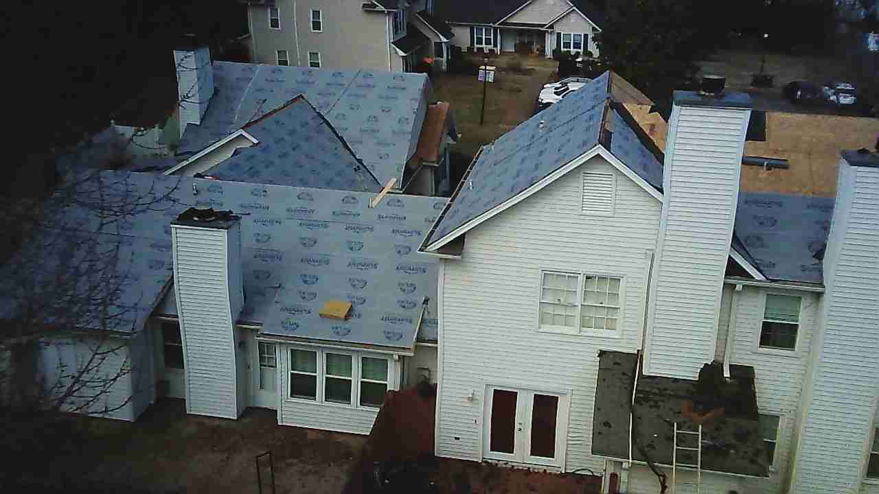 An aerial view of a white multi-story house undergoing roof renovations, with sections covered in blue protective paper.