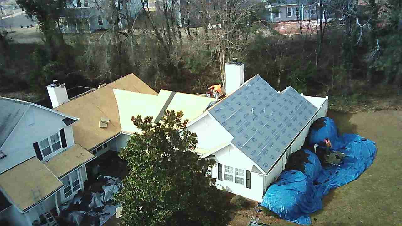 Aerial view of a house undergoing roof repairs, featuring tan and gray shingled sections and a large blue tarp on the lawn.