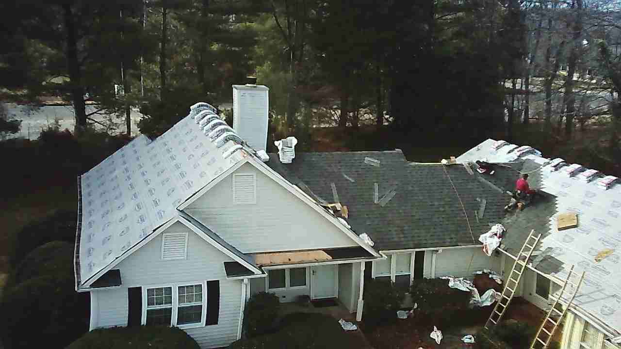 An aerial view of a house undergoing roof repairs, with sections covered in white underlayment and a worker on the roof.