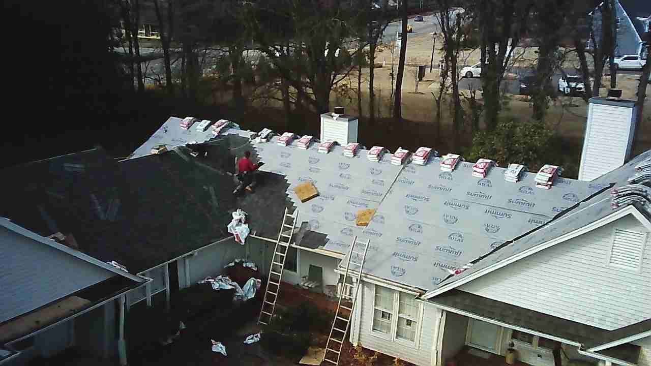 A worker on a ladder performs repairs on a house roof partially covered with grey underlayment and new shingles.