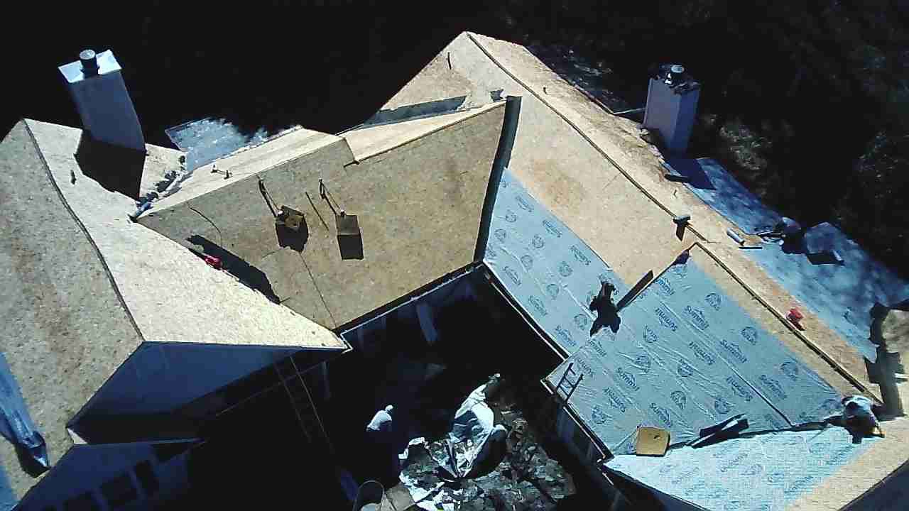 An overhead aerial view of a house roof under construction, showing plywood sheathing and blue underlayment.