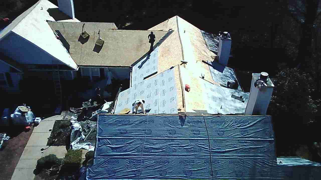 An aerial view of a house roof undergoing construction with workers installing underlayment and shingles.