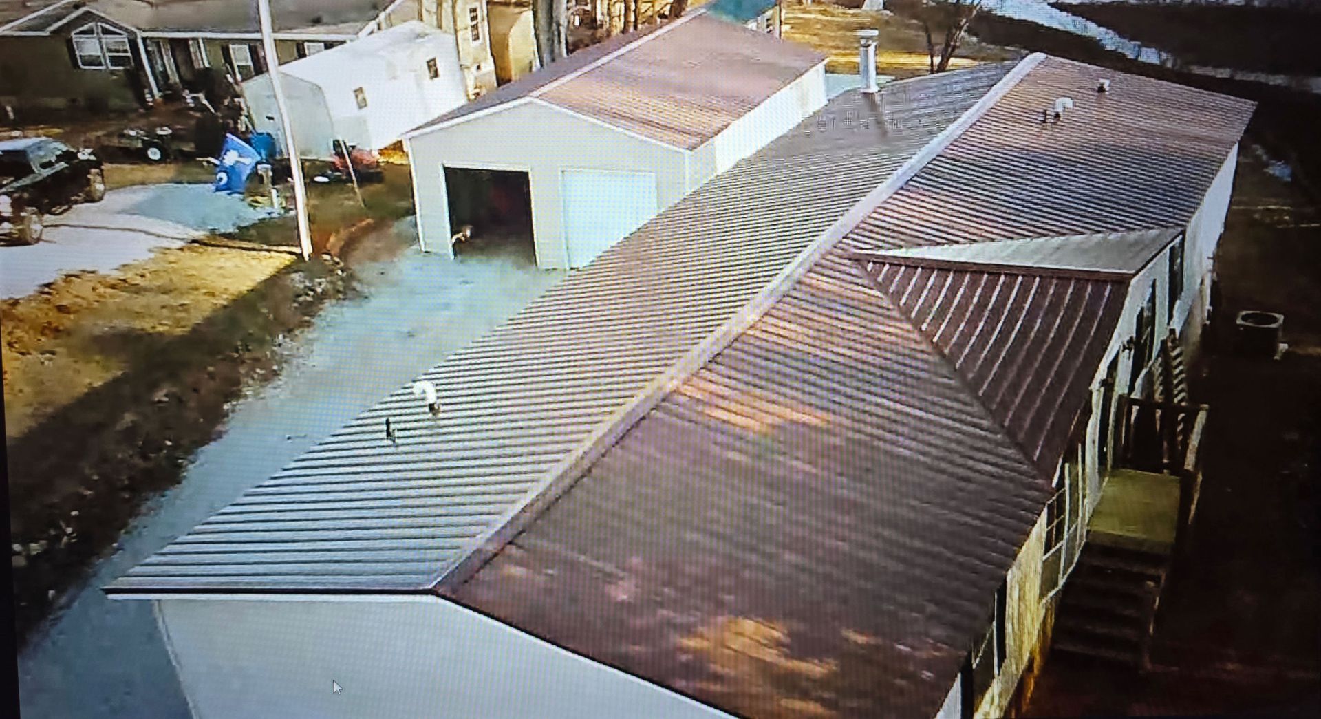 An elevated, angled view of a house with a dark metal roof next to a white detached garage in a residential area.