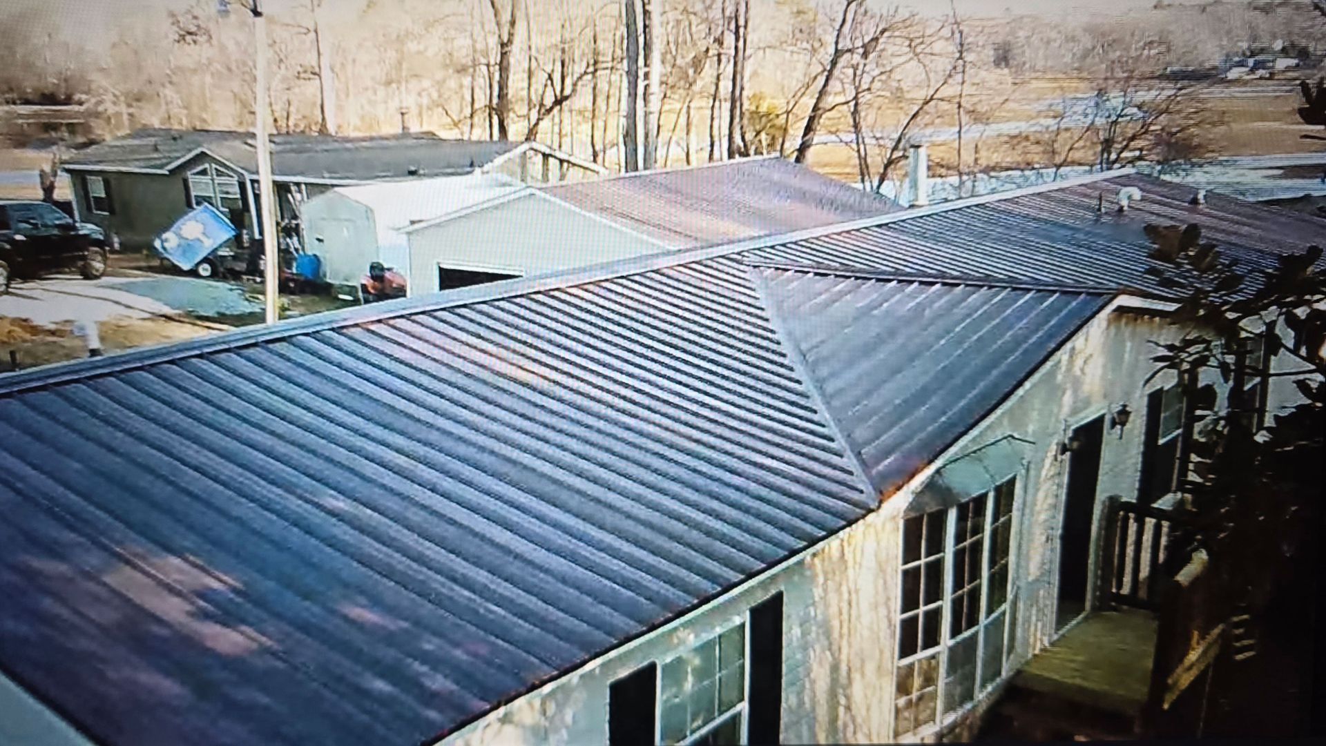 An elevated view of a manufactured home with a dark, ridged metal roof, adjacent to a smaller outbuilding.