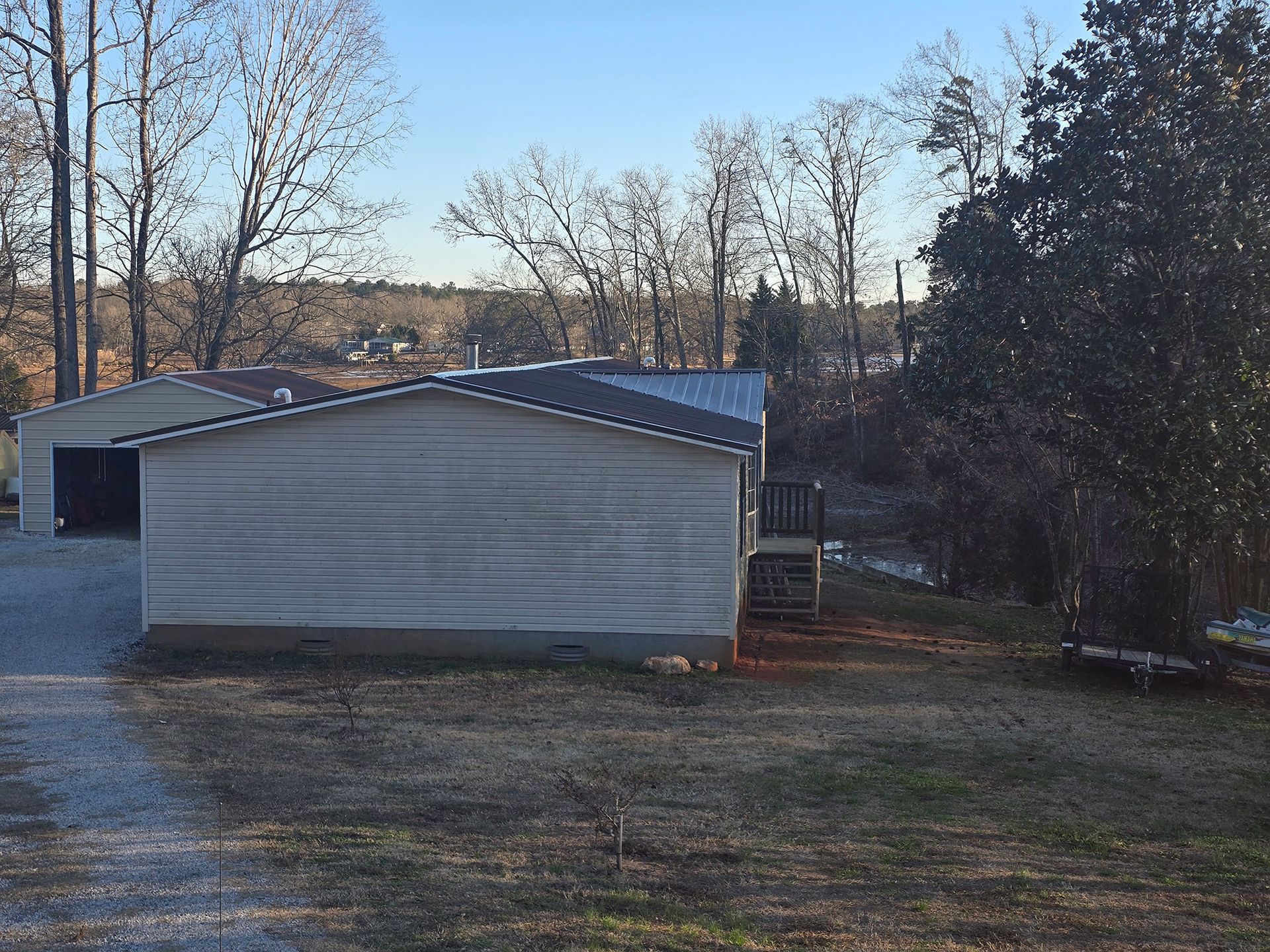A light-colored, single-story house with a metal roof and an attached garage, situated on a grassy lot with trees.