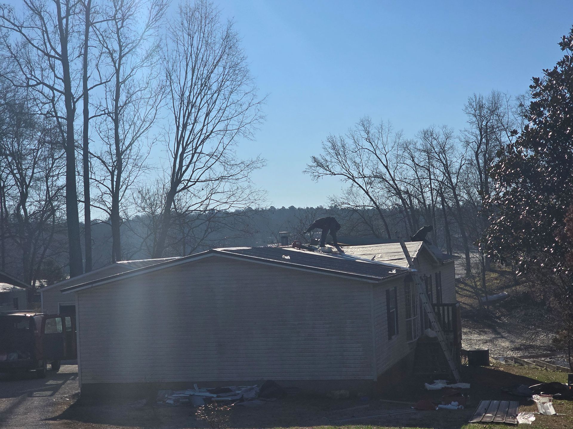 A worker installs metal roofing on a light-colored mobile home surrounded by trees under a clear blue sky.