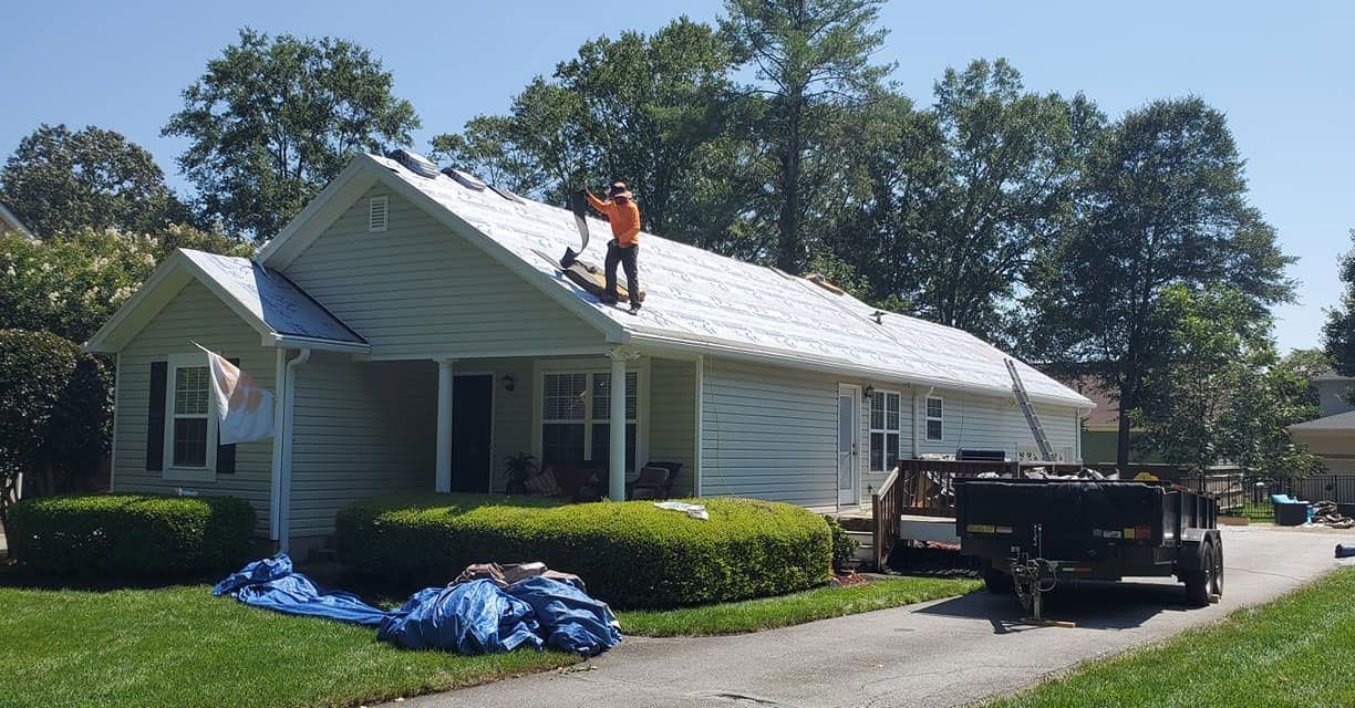 A worker in an orange shirt repairs a house roof covered in white underlayment, with a trailer parked in the driveway.
