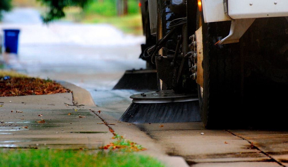 Street sweeper cleaning a sidewalk curb beside a wet road, with green grass and scattered leaves.