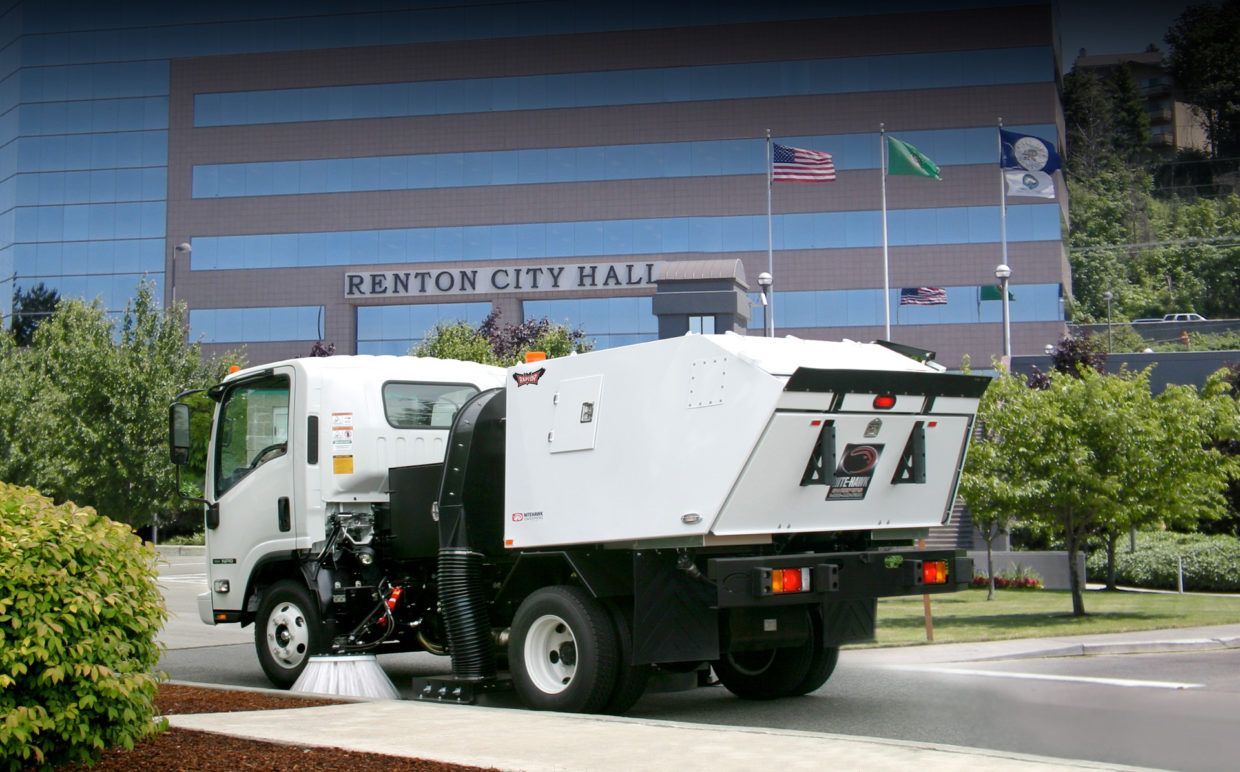 White utility truck driving past Renton City Hall on a sunny street with trees and flags