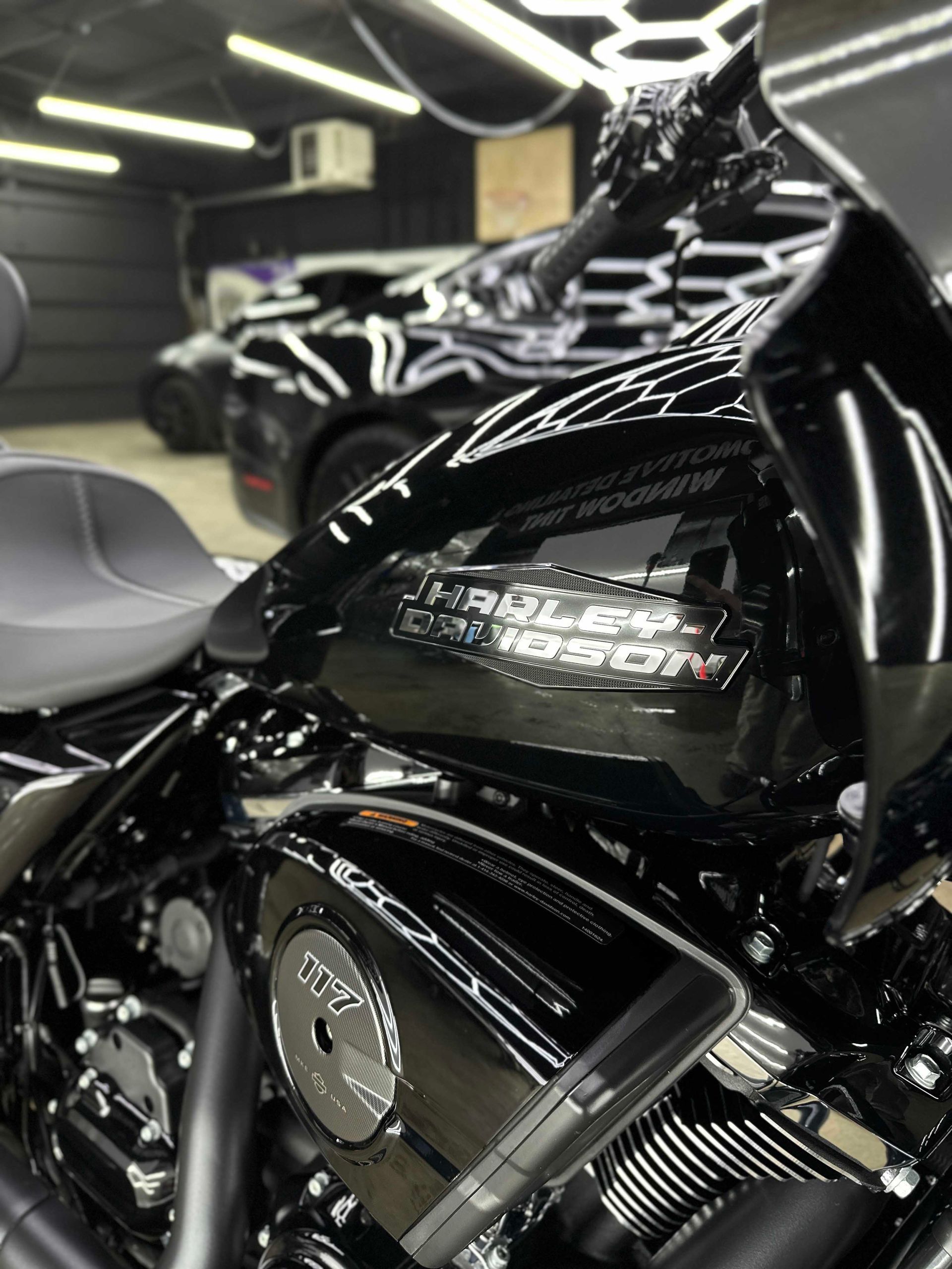 Shiny black Harley-Davidson motorcycle with chrome details, parked indoors; reflection of a car in background.