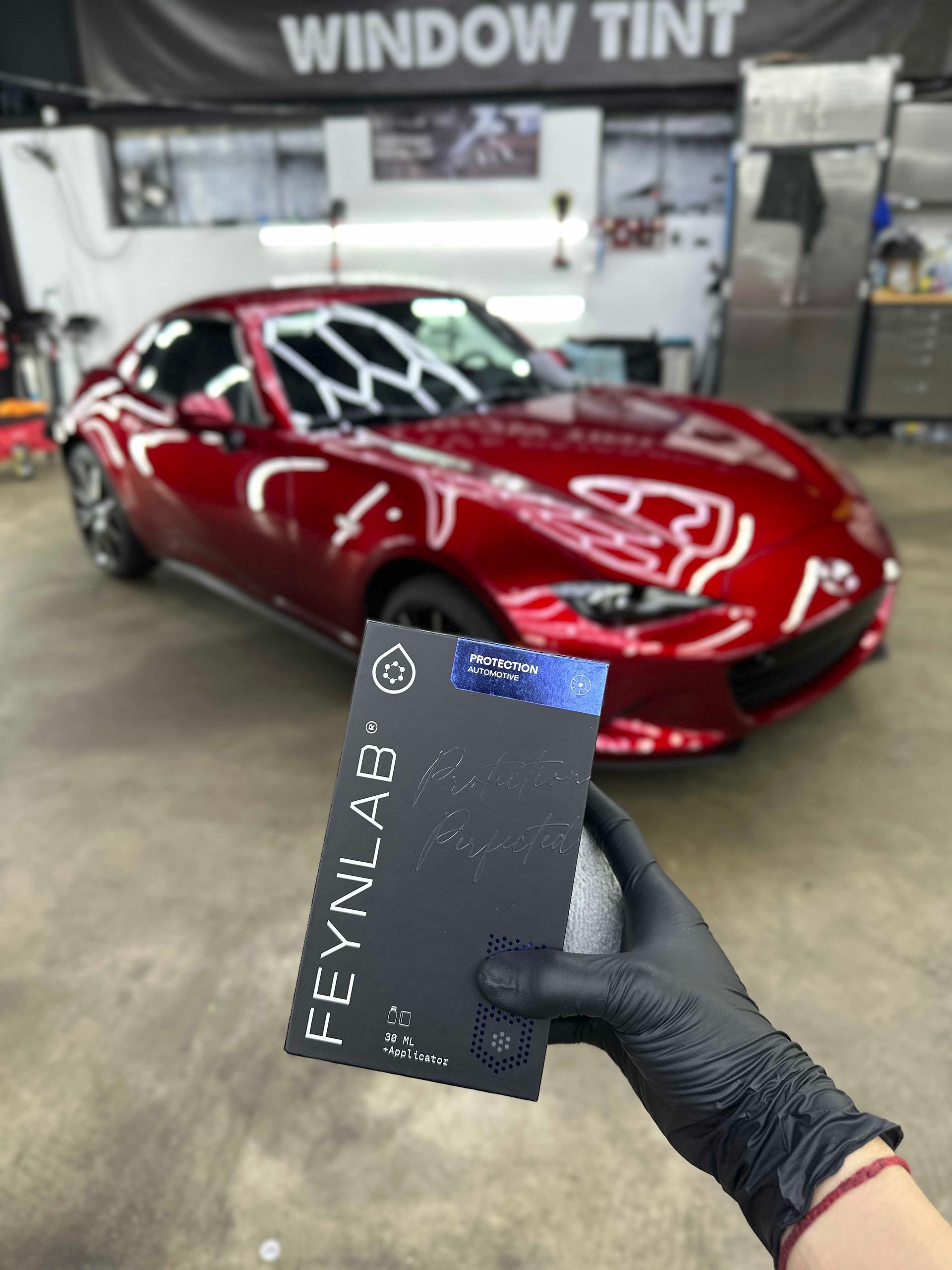 Person in black gloves holds a car protection product box in front of a red Mazda sports car.