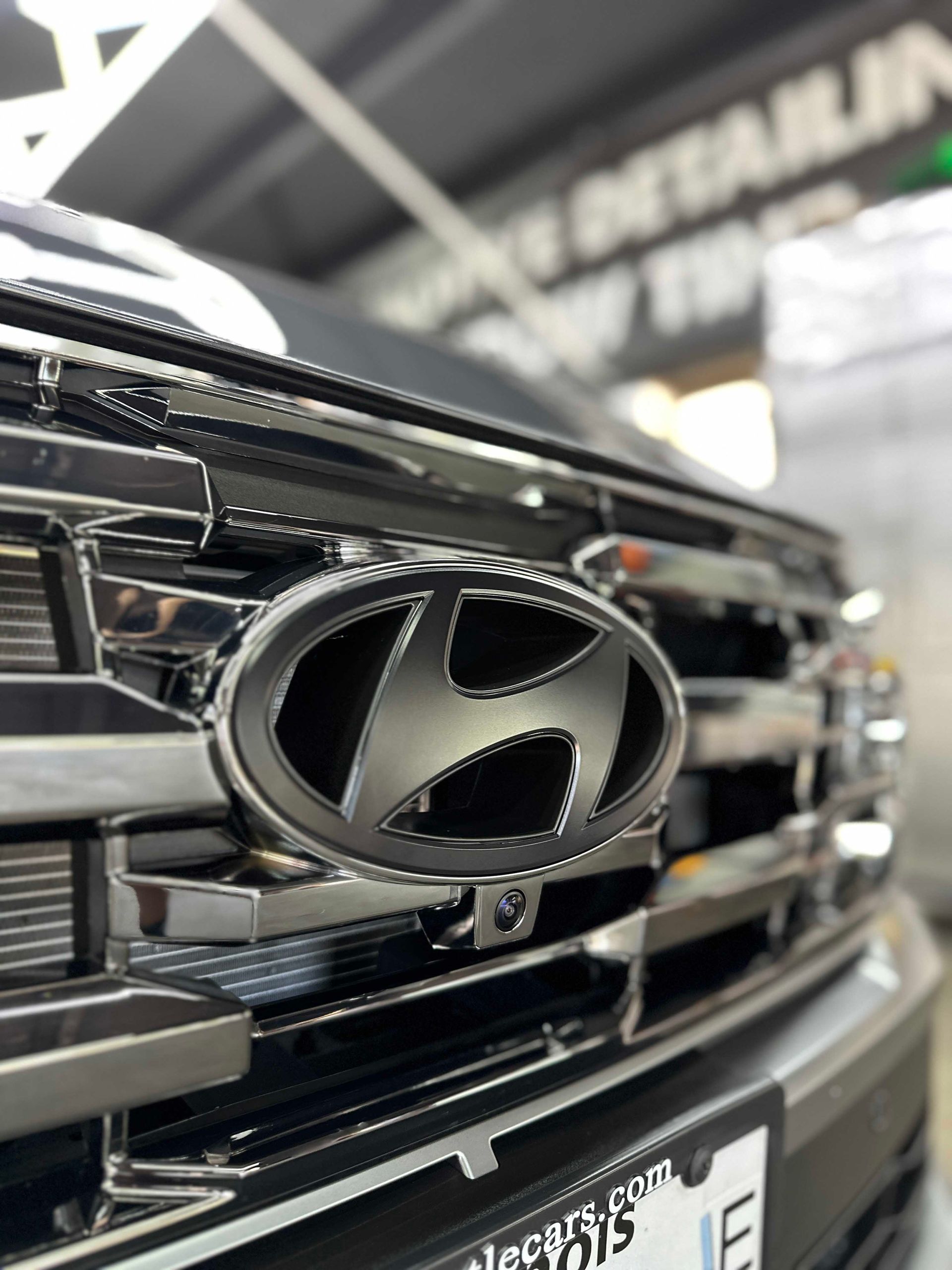 Close-up of a black Hyundai emblem on a chrome grill; vehicle front in a detail shop.