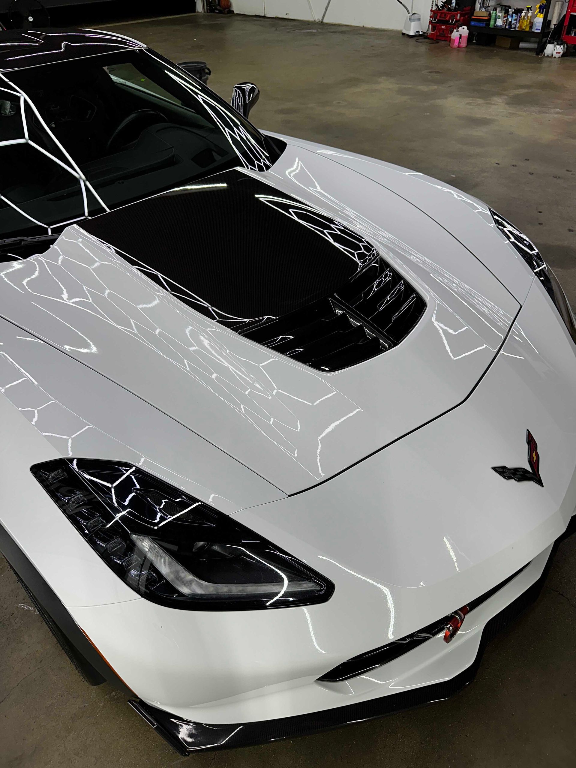 White Chevrolet Corvette with a black hood in a well-lit garage.