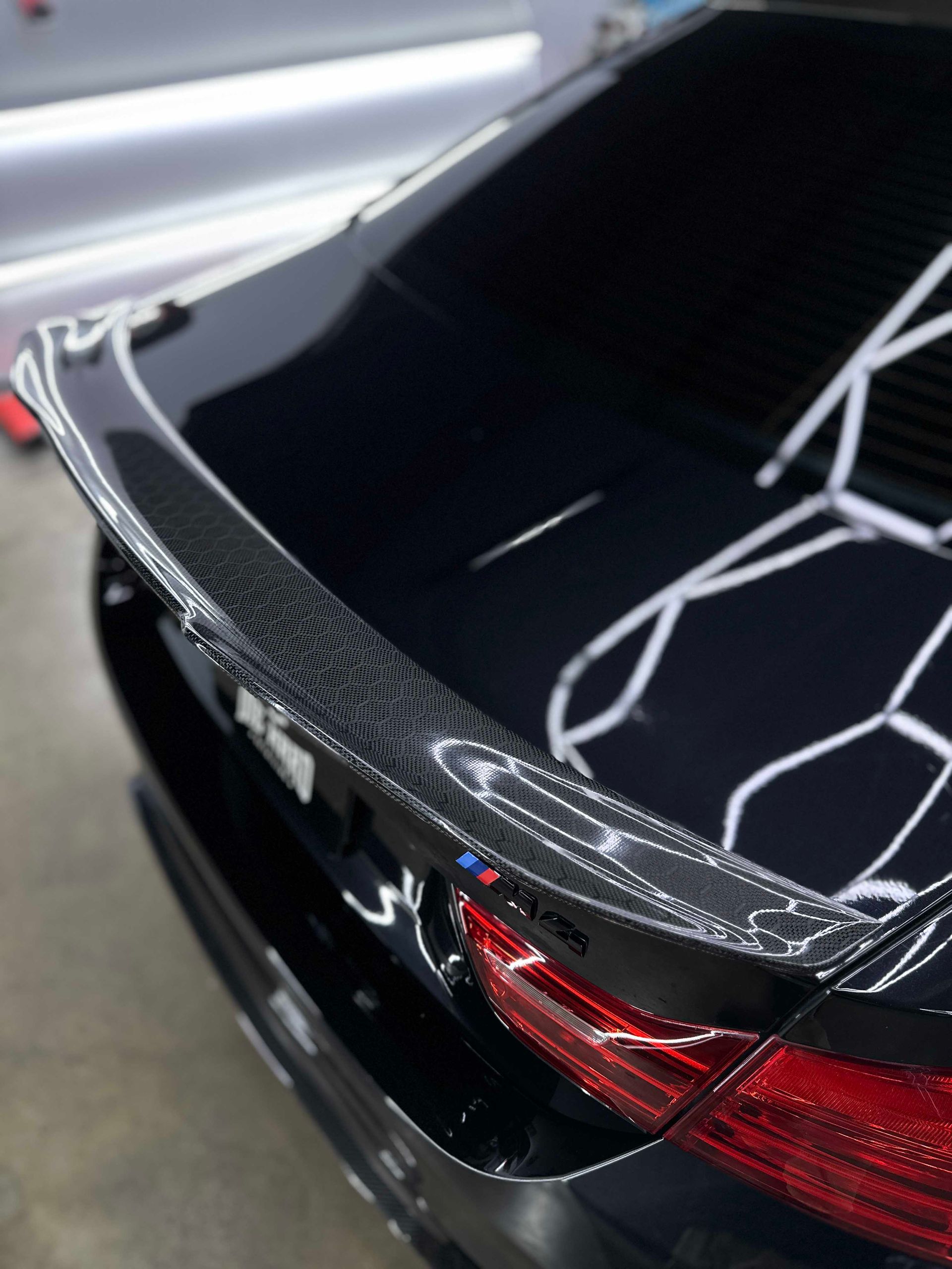 Close-up of a black car's rear with a carbon fiber spoiler. Shiny paint reflects interior lighting.