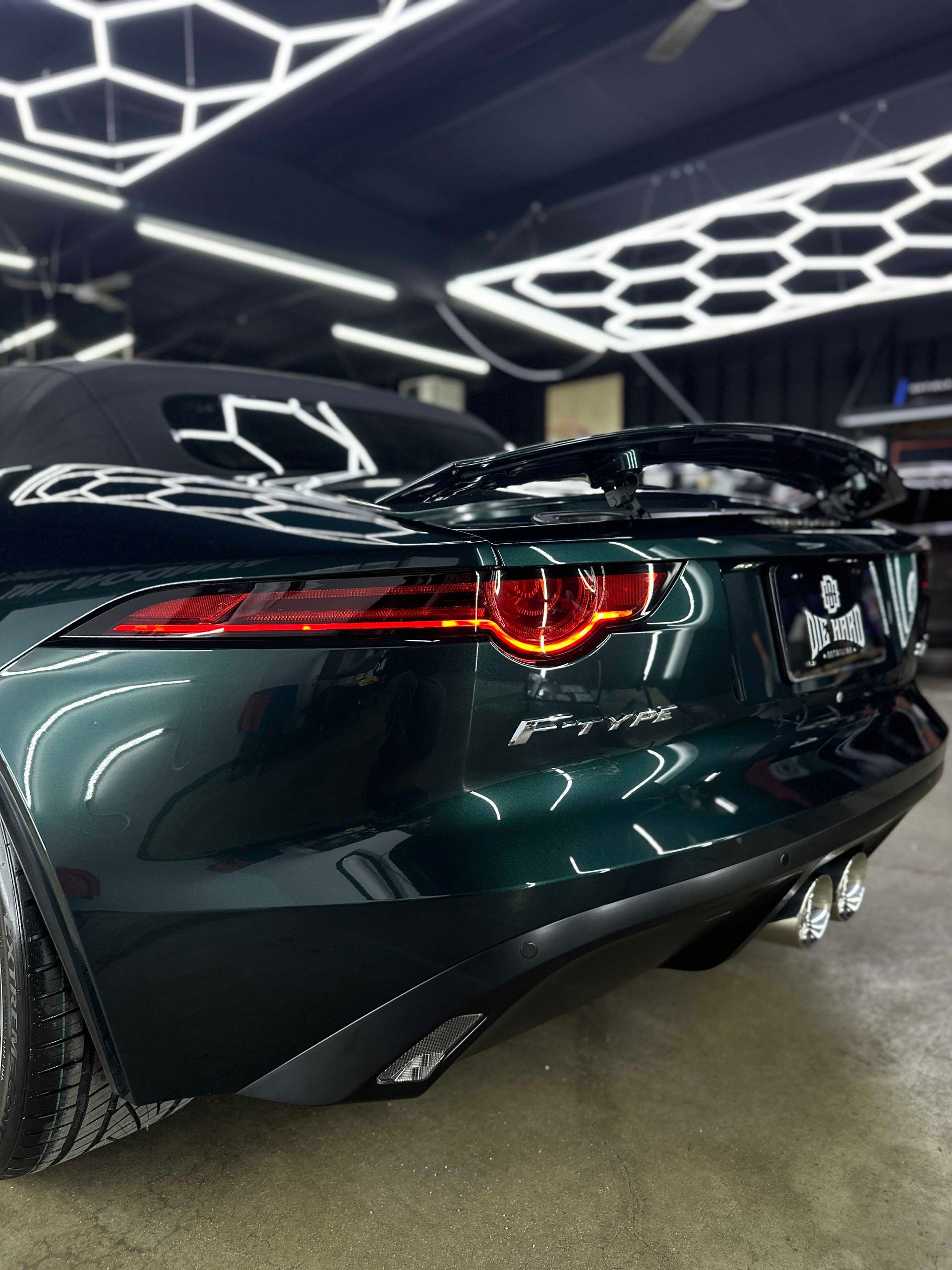 Dark green Jaguar F-Type convertible, rear view, with spoiler, under bright lights.