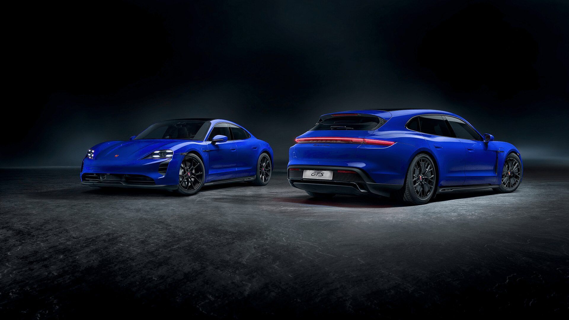 Two blue Porsche Taycans, a sedan and a wagon, on a dark, textured surface.