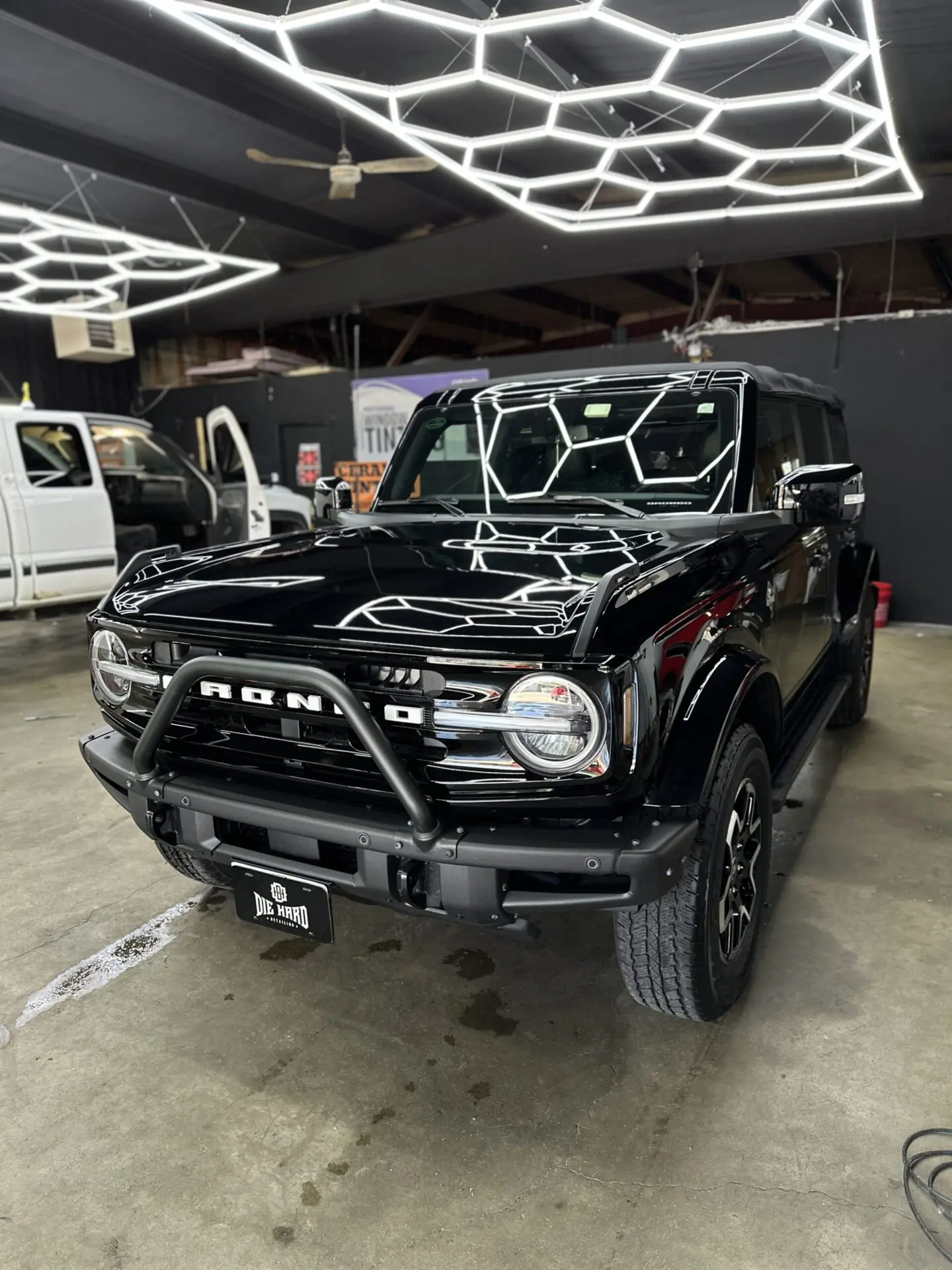 Black Ford Bronco parked in a brightly lit garage, with a protective bar and honeycomb lighting above.