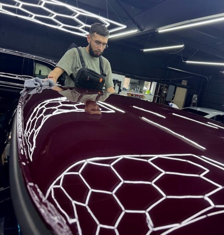 Man polishing a burgundy car roof inside a well-lit garage. Reflective honeycomb lighting is above.
