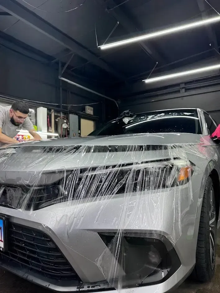 Man applying protective film to a silver car's hood in a garage.